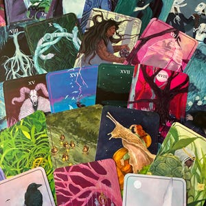 May include: A selection of tarot cards with diverse illustrations. The cards depict a deer, snakes, a woman, a snail, and other mystical figures. The colour scheme includes blues, greens, pinks, and purples.