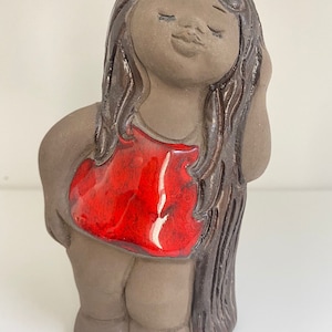 May include: A ceramic figurine of a girl with long brown hair wearing a red dress. The figurine has a simple, rustic style.