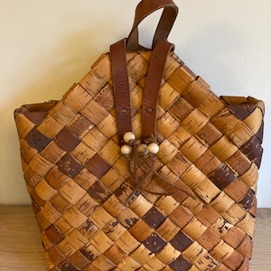May include: A woven birch bark bag with a brown leather strap and wooden beads. The bag is a light brown color with dark brown accents.
