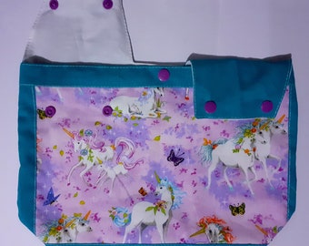 Könnte beinhalten: Eine Tragetasche mit einem Einhorn-Muster auf lavendelfarbenem Hintergrund. Sie hat eine türkisfarbene Umrandung und Klappe, die mit lila Druckknöpfen befestigt ist. Der Stoff zeigt weiße Einhörner mit floralen Mähnen und Schmetterlingen.
