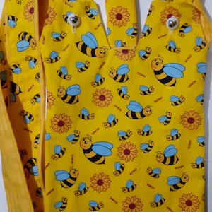 Puede incluir: Bolso tote amarillo con un patrón repetido de abejas de dibujos animados, girasoles y la palabra "HONEY". El bolso tiene una abertura en forma de cuello en V y dos correas. El bolso está hecho de tela amarilla brillante.