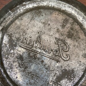 May include: Close-up of the base of a metal container with a worn, aged look. The container is circular with a raised edge. The word "Ruinart" is embossed in a decorative font, suggesting a vintage item.