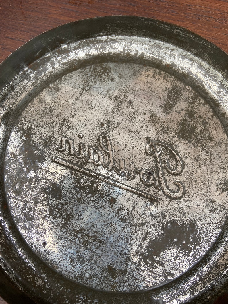 May include: Close-up of the base of a metal container with a worn, aged look. The container is circular with a raised edge. The word "Ruinart" is embossed in a decorative font, suggesting a vintage item.