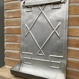 Puede incluir: Un buzón de pared vintage de metal con un patrón de diamante y una bandeja debajo. El buzón está hecho de metal plateado y tiene un aspecto desgastado y rústico.