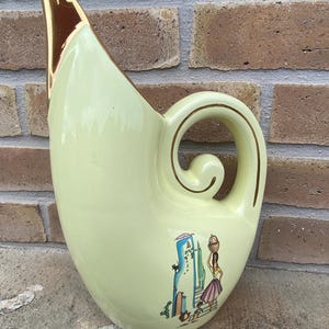 Peut inclure: Pichet en céramique jaune pâle avec un bec verseur incurvé et une anse décorative. Le pichet a un bord et des accents de poignée dorés. Une scène peinte d'une femme portant un panier est sur le devant. Le pichet est probablement vintage.