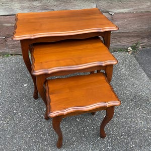 Op de afbeelding: Een set van drie houten nesttafels met een lichtbruine afwerking. De tafels hebben een gegolfde rand en gebogen poten.