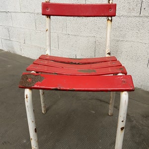 May include: A red metal chair with white painted legs. The chair has a slatted seat and back and shows signs of rust and wear.