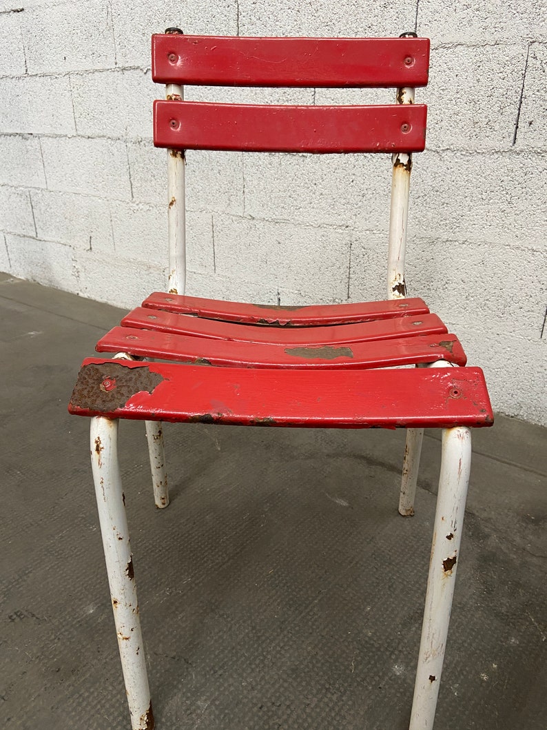 May include: A red metal chair with white painted legs. The chair has a slatted seat and back and shows signs of rust and wear.