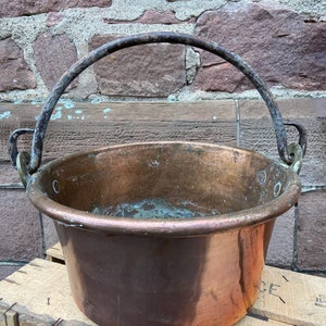 Puede incluir: Una olla grande de cobre con un fondo grueso y redondeado y una robusta asa de hierro. La olla tiene una pátina desgastada y envejecida y está sentada sobre una caja de madera.