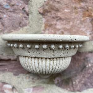 Peut inclure: Un petit pot mural décoratif beige avec un bord perlé et un motif cannelé. Le pot est fait d'un matériau texturé qui ressemble à de la pierre.