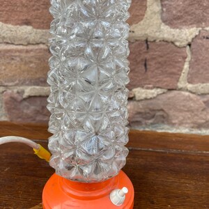 May include: A vintage table lamp with a clear glass shade featuring a geometric pattern. The base is a bright orange color and has a white on/off switch.
