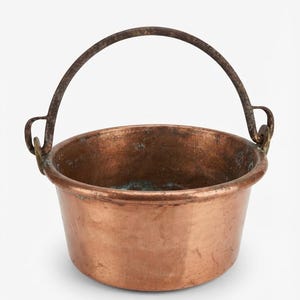 Puede incluir: Una olla redonda de cobre con un asa móvil de metal oscuro y desgastado sujeta a dos anillas laterales. La superficie exterior muestra un tono cobrizo cálido con signos de antigüedad, mientras que el interior presenta una pátina verdosa.