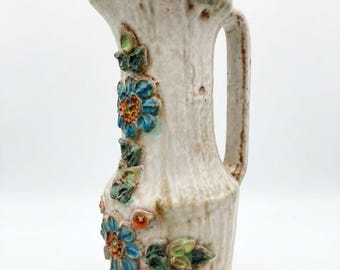 May include: A ceramic pitcher with a textured, off-white glaze and brown vertical streaks. The exterior is decorated with raised, hand-painted floral motifs in shades of blue, green, and orange, featuring a side handle and a pouring spout.