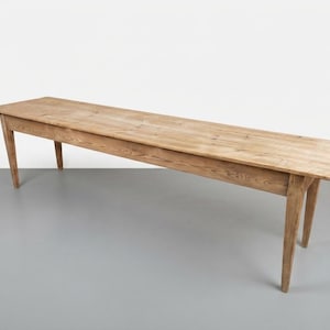 Op de afbeelding: Een lange, rechthoekige houten tafel met een natuurlijke lichtbruine houtnerfafwerking. De tafel heeft een vlak, glad bovenoppervlak en wordt ondersteund door vier taps toelopende poten die vanuit de hoeken naar buiten staan.