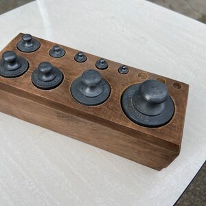 Peut inclure: Un ensemble de poids en fonte vintage dans une boîte en bois. Les poids sont de différentes tailles et ont une finition noire. La boîte est en bois foncé et porte une étiquette indiquant "Set of Weights".
