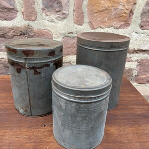 May include: Three antique metal storage tins with lids. The tins are different sizes and show signs of wear, with rust and age marks. They are placed on a wooden surface against a brick wall. The tins are cylindrical and have a rustic, industrial appearance.