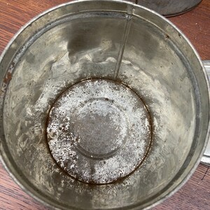 May include: An aged metal container, cylindrical in shape, showing signs of wear and rust inside. The container is placed on a wooden surface, with another similar container in the background. The metal has a dull, aged appearance, suggesting its vintage.