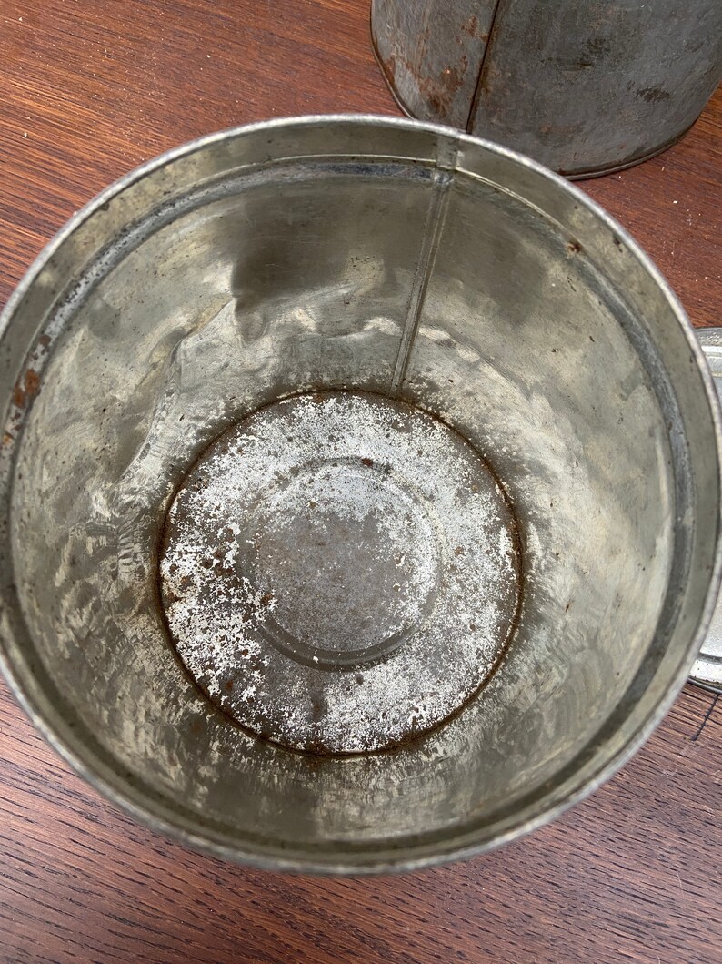 May include: An aged metal container, cylindrical in shape, showing signs of wear and rust inside. The container is placed on a wooden surface, with another similar container in the background. The metal has a dull, aged appearance, suggesting its vintage.