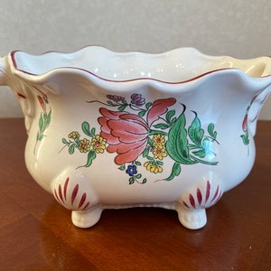 Pode incluir: Um vaso de cerâmica branco com uma borda ondulada e três pés. O vaso é decorado com um desenho floral em tons de vermelho, amarelo, verde e azul.