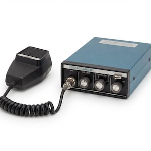 Op de afbeelding: Een blauwe Zodiac mini-6 radio-ontvanger met een zwarte microfoon verbonden door een spiraalsnoer. Het voorpaneel bevat drie zwarte knoppen met zilveren doppen, een microfooningang en de tekst ZODIAC mini-6, MIC, VOLUME, SQUELCH en SG.