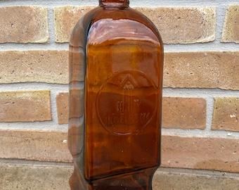 May include: A tall, amber-coloured glass bottle with a black cap. The bottle has a square shape with rounded edges and a raised base. The bottle has a circular label with the text "The J.B. Roellig Co."