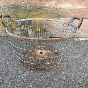 Puede incluir: Una cesta redonda de alambre metálico antigua con dos asas arqueadas. La cesta tiene un aspecto oxidado y envejecido, con una base circular y una estructura en forma de jaula. Adecuada para almacenamiento o exhibición.