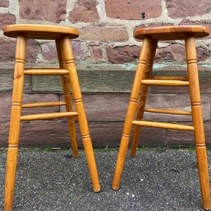 May include: Two wooden bar stools with a natural finish. The stools have a simple design with four legs and a round seat.