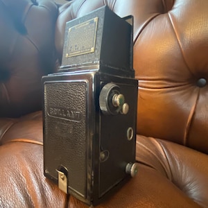May include: A vintage black and silver camera with the word "BRILLANT" embossed on the front. The camera has a textured leather-like finish and several dials and knobs. A small metal plate is affixed to the top. The camera is resting on a brown leather surface.