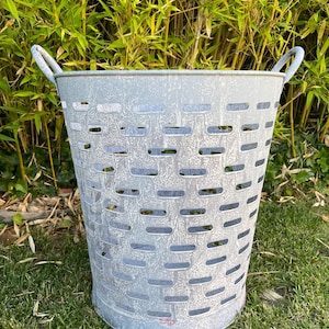 Op de afbeelding: Een witte metalen emmer met een verweerde afwerking en een geweven patroon. De emmer heeft twee handvatten en staat op een bed van groen gras.
