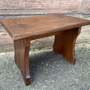 May include: A dark brown wooden stool with a simple design. The stool has a rectangular top and two legs on each side. The legs are slightly curved and have a simple, rounded shape.
