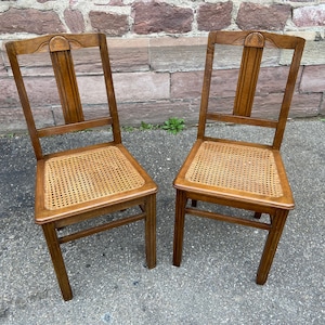 Könnte beinhalten: Zwei Holzstühle mit Rohrgeflechtsitzen. Die Stühle haben ein schlichtes Design mit einer abgerundeten Oberseite und geraden Beinen. Die Stühle sind in gutem Zustand und wären eine großartige Ergänzung für jedes Zuhause.