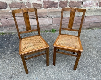 Könnte beinhalten: Zwei Holzstühle mit Rohrgeflechtsitzen. Die Stühle haben ein schlichtes Design mit einer abgerundeten Oberseite und geraden Beinen. Die Stühle sind in gutem Zustand und wären eine großartige Ergänzung für jedes Zuhause.
