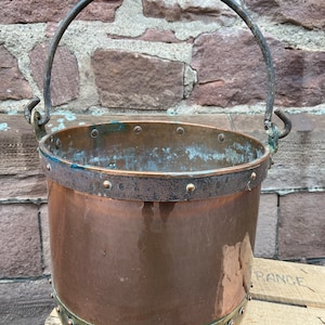 May include: A large copper bucket with a metal handle and rivets. The bucket is sitting on a wooden crate.