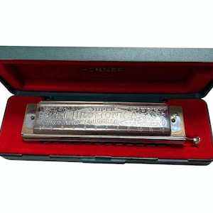 Könnte beinhalten: Eine silberne Super Chromonica Mundharmonika in einem grünen Etui mit rotem Samtinnenraum. Auf der Mundharmonika steht "Super Chromonica". Das Etui ist geöffnet und zeigt das Instrument.
