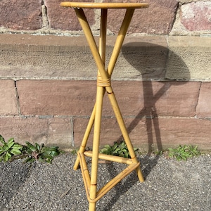 May include: A natural woven bamboo plant stand with a round top and a triangular base. The stand is light brown in colour and has a rustic, vintage look.