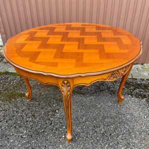 Peut inclure: Une table ronde en bois avec une finition marron clair et un dessus à motifs. La table a des pieds incurvés et des sculptures décoratives. Le diamètre de la table est d'environ 122 cm.