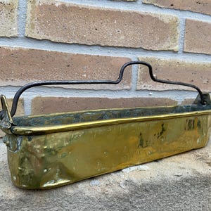 Peut inclure: Un pot en laiton avec une poignée en métal. Le pot est rectangulaire et a un fond légèrement arrondi. La poignée est fixée sur les côtés du pot et est incurvée.