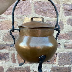 May include: A large copper pot with a lid and a black iron handle. The pot has three black iron legs and is sitting on a brick wall.