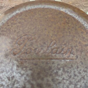 May include: Close-up of a weathered, circular metal object with the embossed word "Pourlair". The surface has a mottled, rust-like appearance, suggesting age and exposure to the elements. The text is in a cursive font.