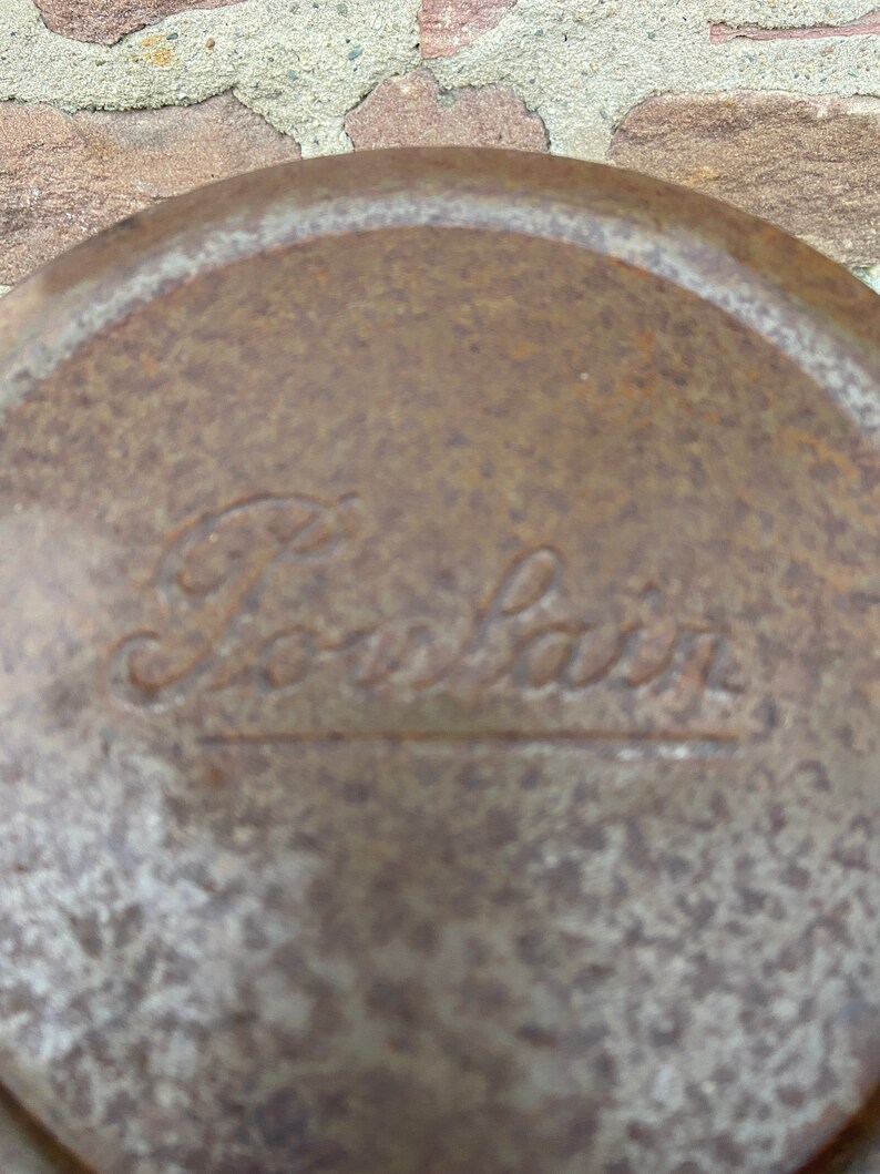 May include: Close-up of a weathered, circular metal object with the embossed word "Pourlair". The surface has a mottled, rust-like appearance, suggesting age and exposure to the elements. The text is in a cursive font.