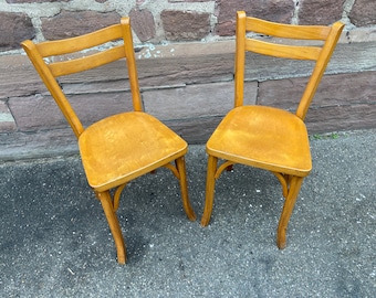 May include: Two wooden chairs with a light brown finish. The chairs have a simple design with a curved back and four legs.