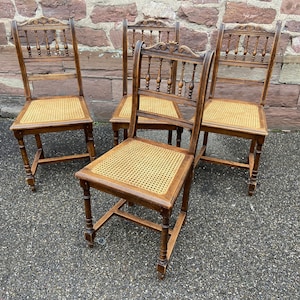Op de afbeelding: Vier antieke houten stoelen met rieten zittingen. De stoelen hebben een donkerbruine afwerking en ingewikkelde houtsnijwerk op de rugleuningen.