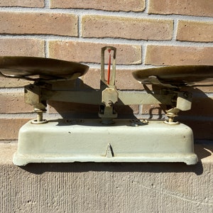 Peut inclure: Une ancienne balance de cuisine avec deux plateaux en bronze et un indicateur central. La balance a une base et un cadre vert clair, avec une aiguille rouge. Le fond est un mur de briques.