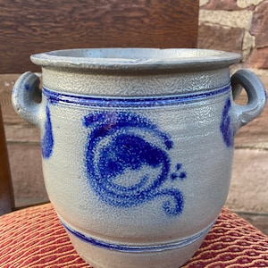 May include: A grey stoneware crock with blue painted designs. The crock has two handles and a wide opening.