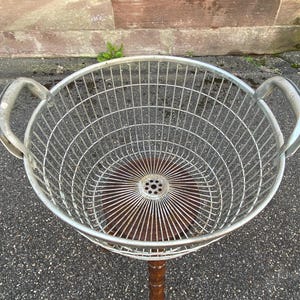 Puede incluir: Una cesta de alambre de metal vintage con dos asas curvas. La cesta tiene una base de madera y está sostenida por un soporte de madera. El diseño de alambre de la cesta crea un patrón visualmente atractivo.