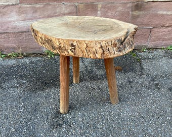 Puede incluir: Un taburete de madera rústico con una tapa redonda en forma de tronco de árbol y tres patas. El taburete está hecho de madera natural con una textura rugosa similar a la corteza.