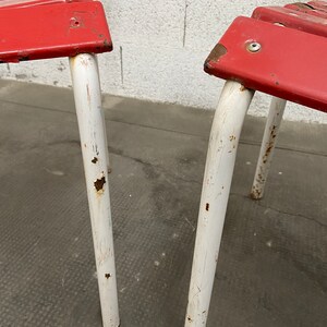 May include: Two red metal chairs with white painted legs. The chairs have a simple design and show signs of wear and rust.