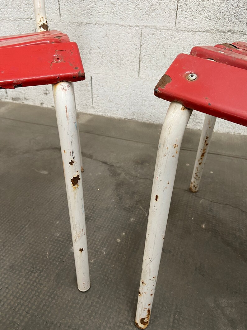 May include: Two red metal chairs with white painted legs. The chairs have a simple design and show signs of wear and rust.