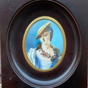May include: A framed oval portrait of a woman wearing a straw hat and a white dress with a floral garland. The portrait is painted in watercolor and gouache on ivory. The frame is made of dark wood.