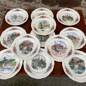 May include: A collection of ten cream-colored ceramic plates with scalloped edges, each featuring a unique, colorful pastoral scene in the center and decorative floral motifs in red, blue, and green along the rim of the dinnerware set.